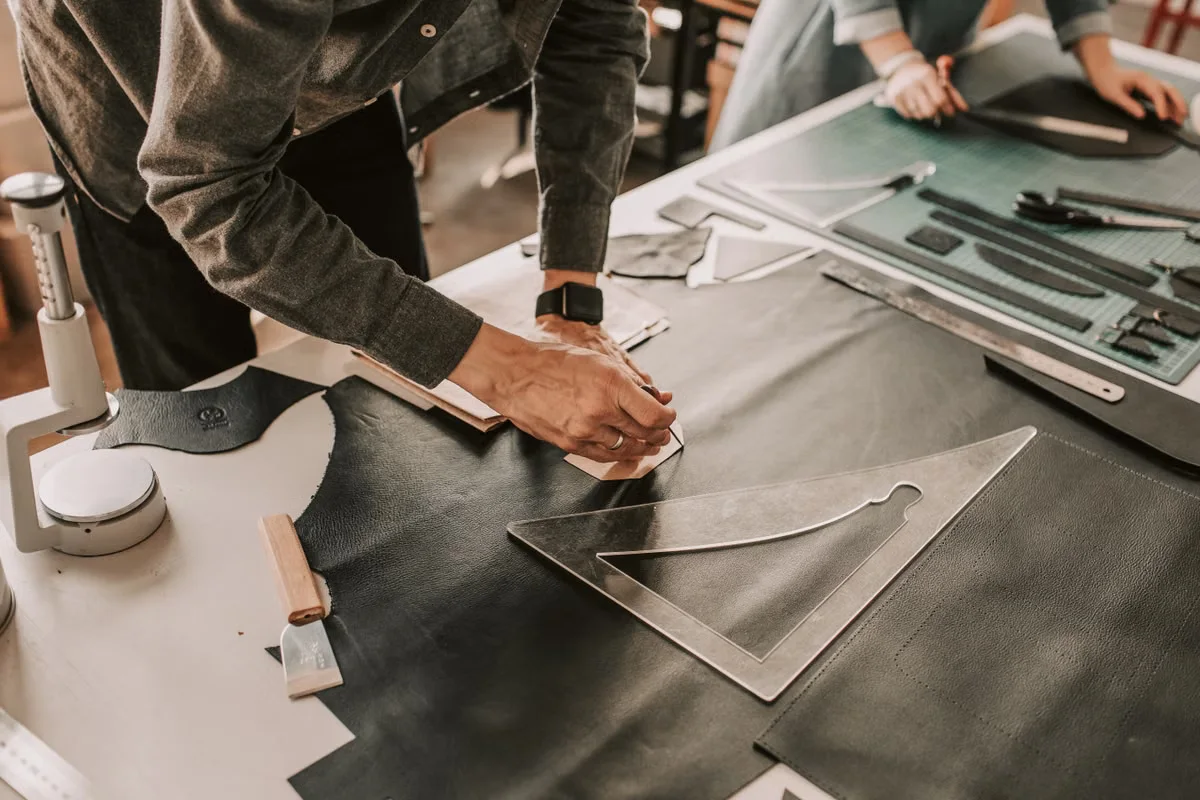 Two leather craftsmen working side by side at a workshop bench, hand-cutting large panels of dark leather with precision tools — the kind of skilled artisan workforce that distinguishes a serious leather goods manufacturer