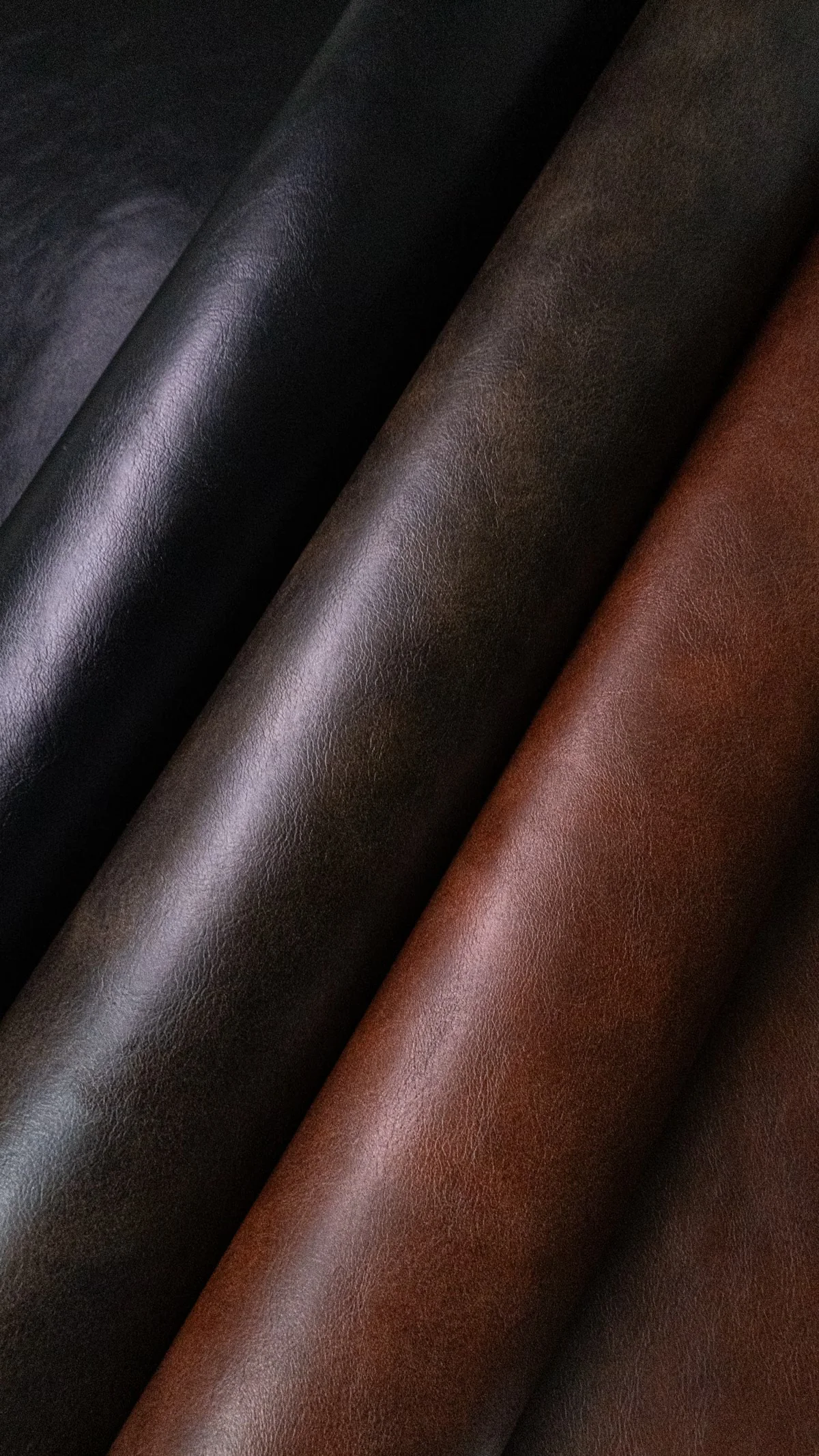 Multiple rolls of smooth leather hide in black, dark brown, and cognac displayed side by side, showing the color range and surface finish of full-grain and top-grain leather grades available for wholesale bag production