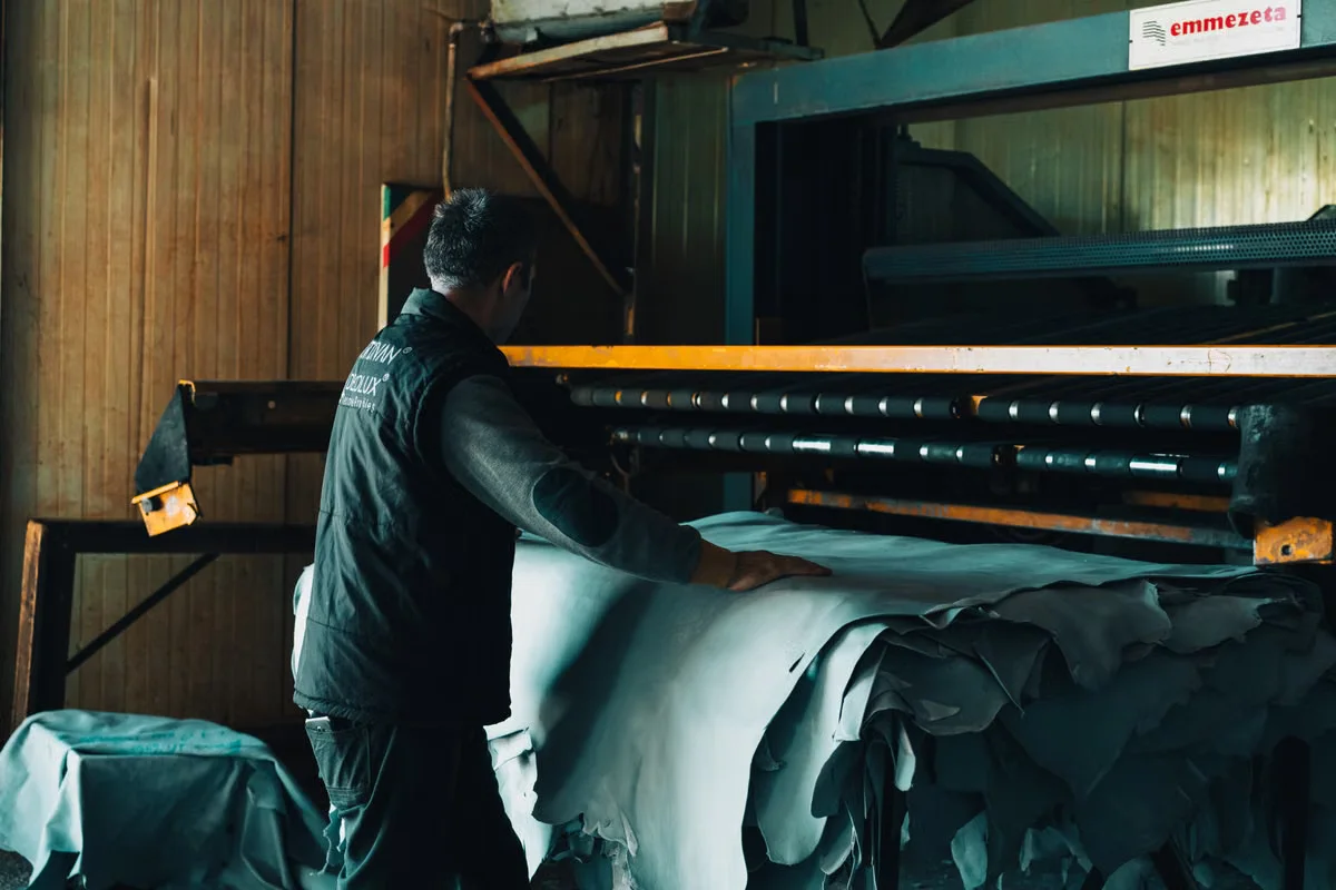 Leather factory worker laying out and inspecting large sheets of tanned hide at an industrial processing machine — the raw material stage that drives minimum order requirements in leather bag manufacturing