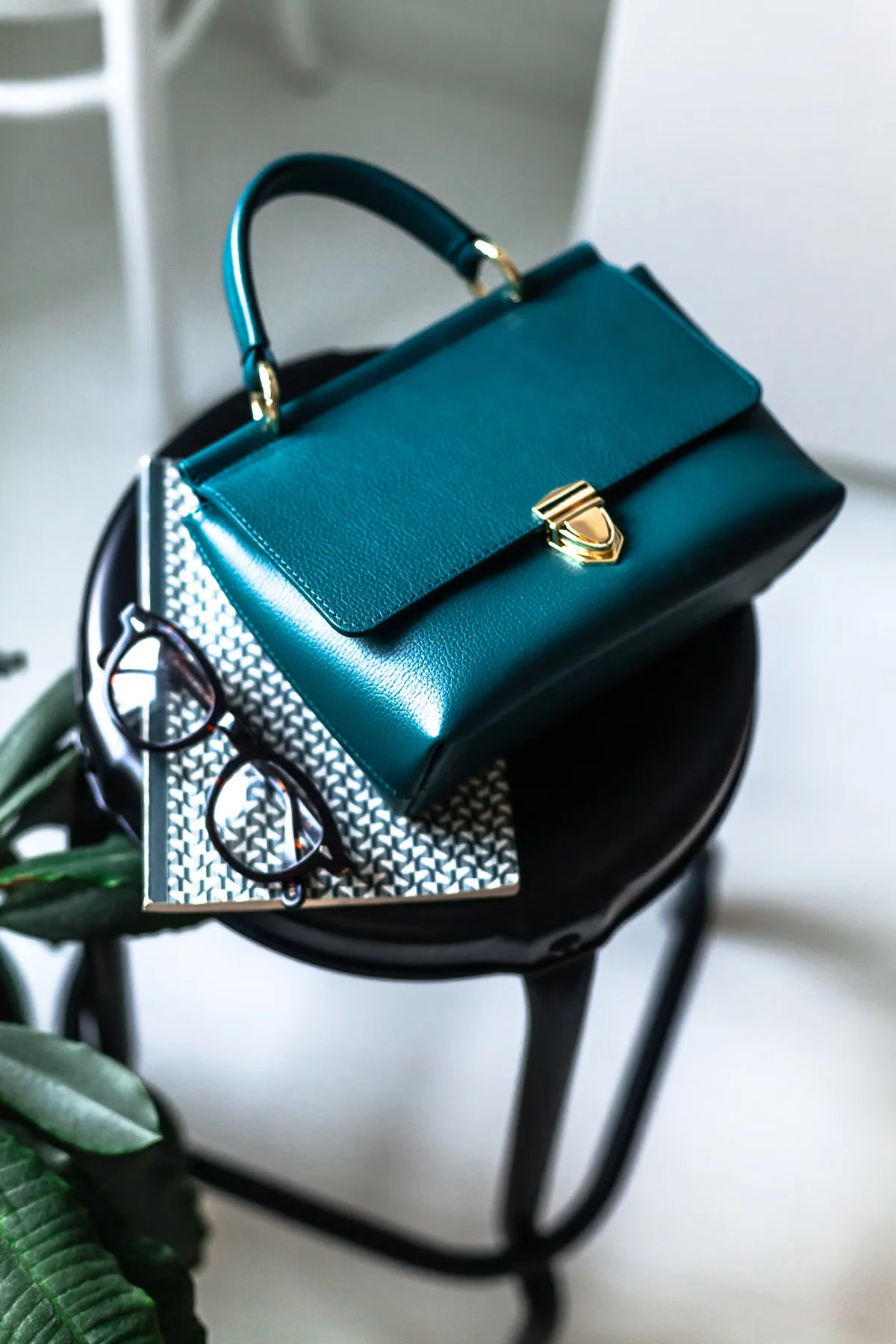 A structured teal leather top-handle bag resting on a notebook beside reading glasses — representing the design brief and sample communication stage of the leather bag sourcing process