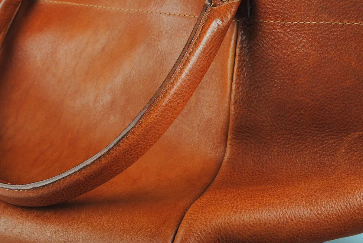 Close-up detail of a tan leather bag showing strap attachment, pebbled grain texture and zipper hardware — the kind of construction detail that must be explicitly specified in a leather bag tech pack to ensure accurate factory production