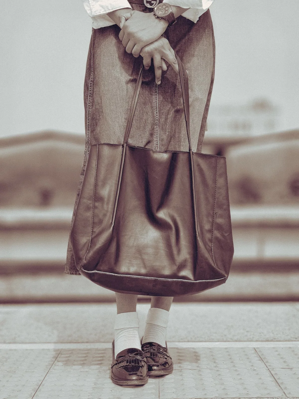 A person holding an oversized dark leather tote bag with both hands at their sides — the bag's proportions relative to the human body illustrate the generous scale of an oversized tote format and how it reads in real-world carry