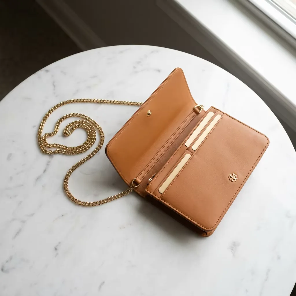 A tan genuine leather wallet on chain opened flat on a marble table, gold chain curled beside it, card slots and interior compartments visible